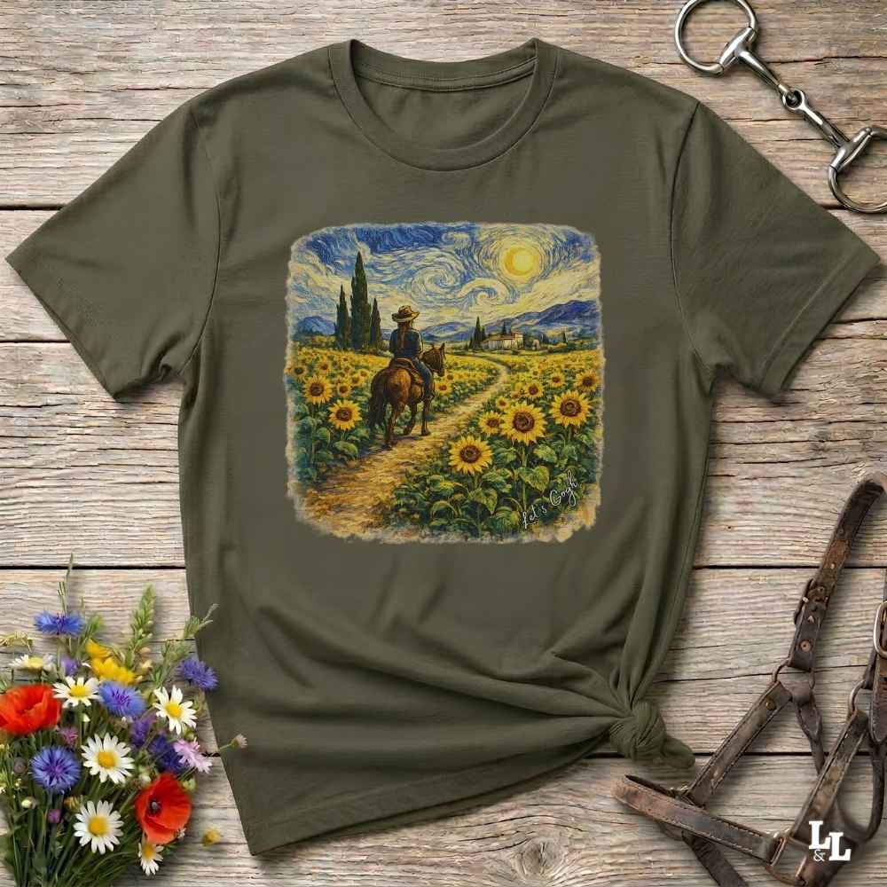 An olive green short-sleeve t-shirt with a square graphic print of a cowboy riding through a sunflower field in a Van Gogh-inspired style. The shirt is styled in a flat-lay on a rustic wooden background with decorative flowers and leather tack.