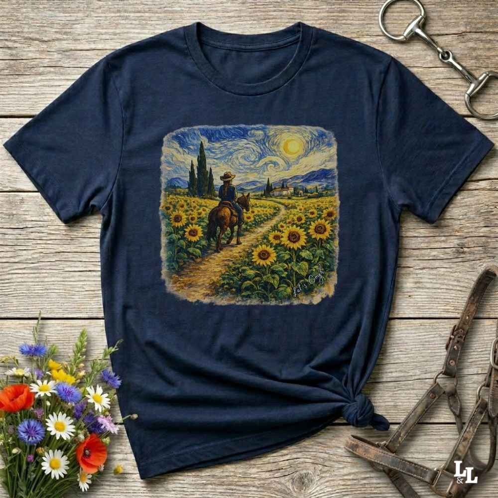 A navy blue graphic t-shirt laid flat on a rustic wooden surface, styled with a bouquet of flowers, a horse bit, and a leather bridle. The t-shirt features a central square print depicting a cowboy riding a horse through a sunflower field under a swirling, impressionistic sky.