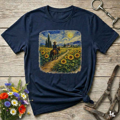A navy blue graphic t-shirt laid flat on a rustic wooden surface, styled with a bouquet of flowers, a horse bit, and a leather bridle. The t-shirt features a central square print depicting a cowboy riding a horse through a sunflower field under a swirling, impressionistic sky.