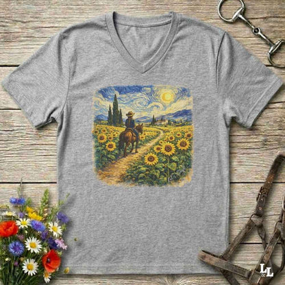 A heather gray v-neck t-shirt laid flat on a rustic wooden surface, featuring a colorful graphic print of a cowboy riding through a sunflower field in a Van Gogh-inspired style. The shirt is styled with equestrian tack, a bouquet of wildflowers, and horse boots.