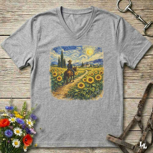 A heather gray v-neck t-shirt laid flat on a rustic wooden surface, featuring a colorful graphic print of a cowboy riding through a sunflower field in a Van Gogh-inspired style. The shirt is styled with equestrian tack, a bouquet of wildflowers, and horse boots.
