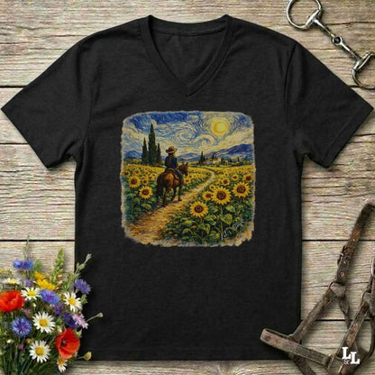 A black V-neck t-shirt laid flat on a rustic wooden surface, featuring a square graphic print of a cowboy riding through a sunflower field in a Van Gogh-inspired style. The shirt is styled with equestrian accessories (reins and a bit) and a small bouquet of wildflowers.