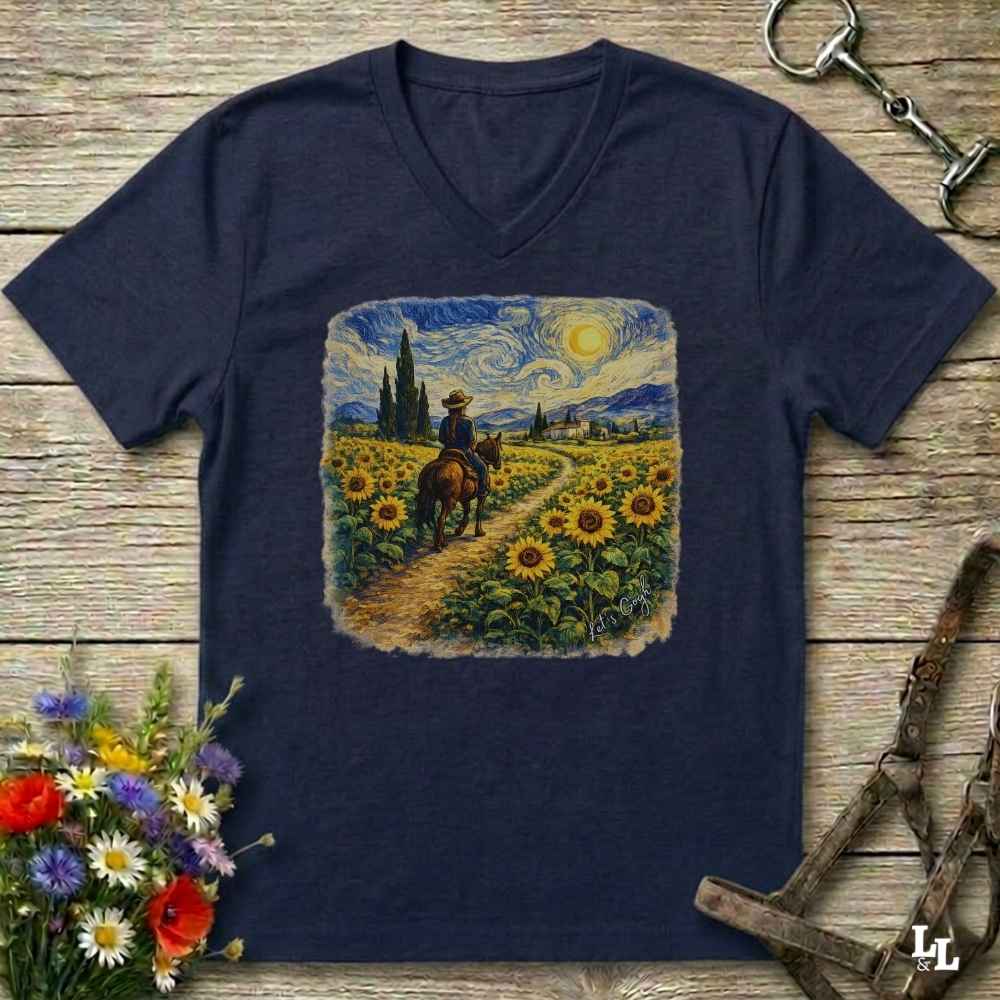 A navy blue V-neck t-shirt laid flat on a rustic wooden surface, featuring a square graphic print of a sunflower field with a rider on horseback in a Van Gogh-inspired style. The shirt is styled with equestrian tack in the background and a small bouquet of wildflowers in the lower left corner.