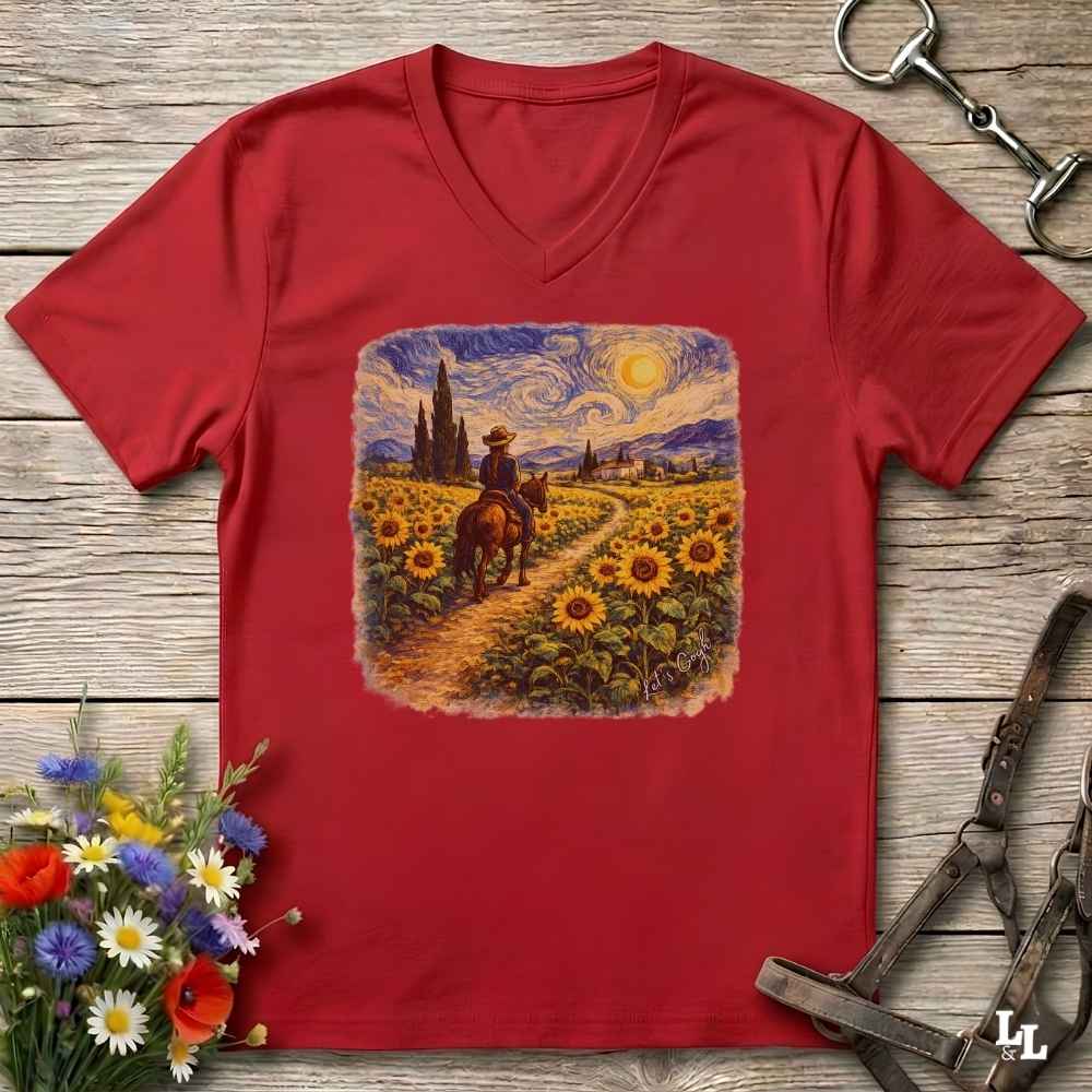 A red V-neck t-shirt laid flat on a rustic wooden surface, featuring a square graphic print of a cowboy riding through a sunflower field in a Van Gogh-inspired style. The shirt is styled with equestrian tack in the top right and a small bouquet of wildflowers in the bottom left.