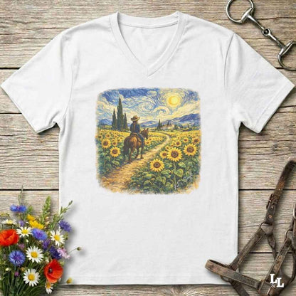 A white V-neck t-shirt laid flat on a rustic wooden surface, featuring a colorful graphic print of a cowboy riding through a sunflower field in a Van Gogh-inspired style. The shirt is styled with equestrian tack, a bouquet of wildflowers, and a horse bit.