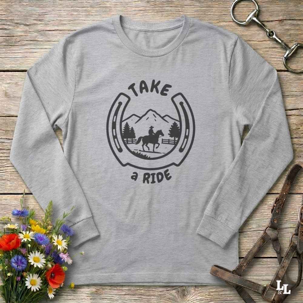A light heather gray long-sleeve t-shirt with a black graphic print reading 'TAKE a RIDE' around a horse and mountain scene inside a horseshoe. The shirt is laid flat on a rustic wooden surface with decorative flowers, a horse bit, and a leather bridle around it.