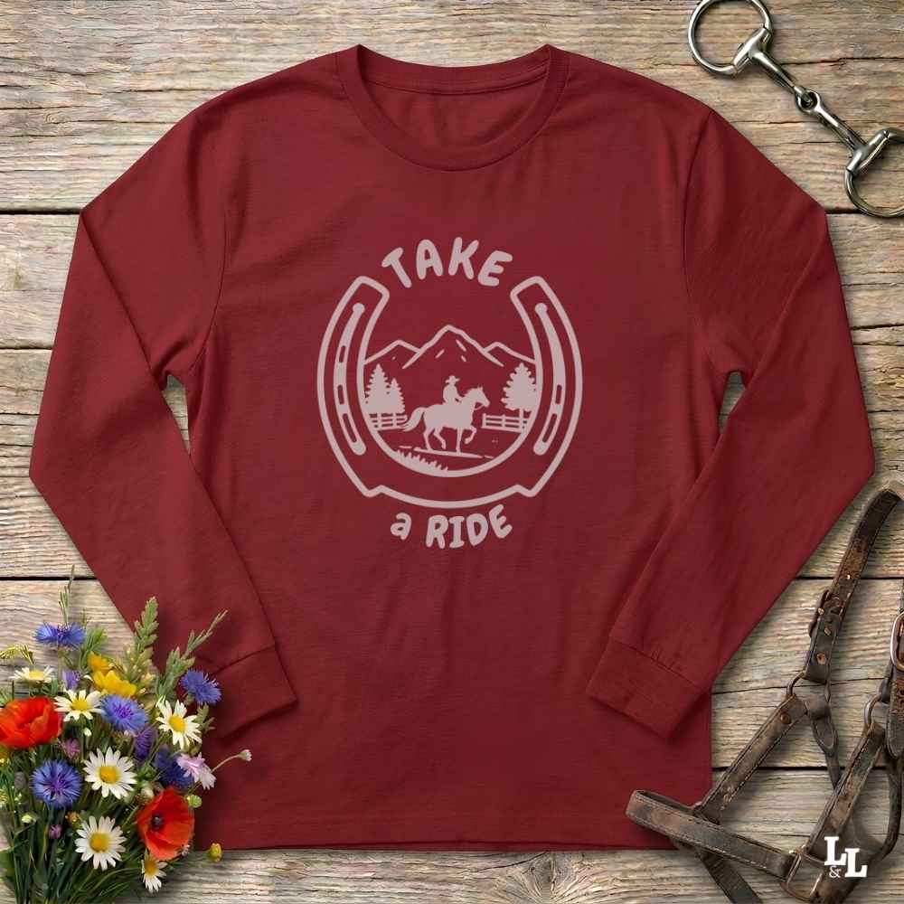 A long-sleeve crewneck t-shirt in maroon red, laid flat on a rustic wooden background. The shirt features a white graphic print in the center showing a horseshoe outline with a horse and mountain design, and the words 'TAKE a RIDE'.