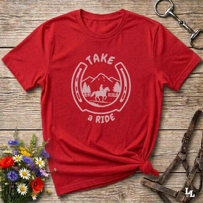 A red short-sleeve graphic T-shirt laid flat on a rustic wooden surface, with a white horseshoe-and-horse design and the words “TAKE a RIDE” printed on the front. The shirt is styled with flowers, a horse bit, and a leather halter nearby.