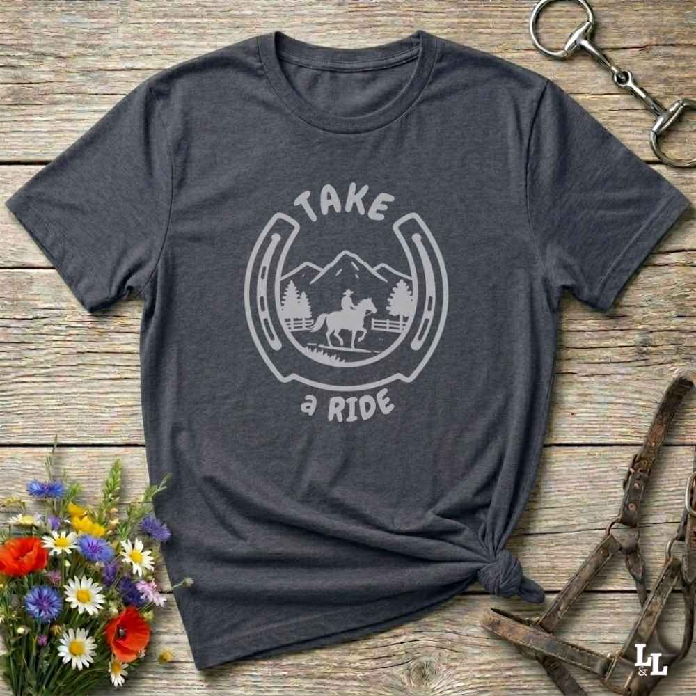 A dark heather gray t-shirt with a white graphic print reading 'TAKE A RIDE' around a horse and mountain scene inside a horseshoe. The shirt is styled in a flat lay on a rustic wooden background with equestrian accessories and flowers.
