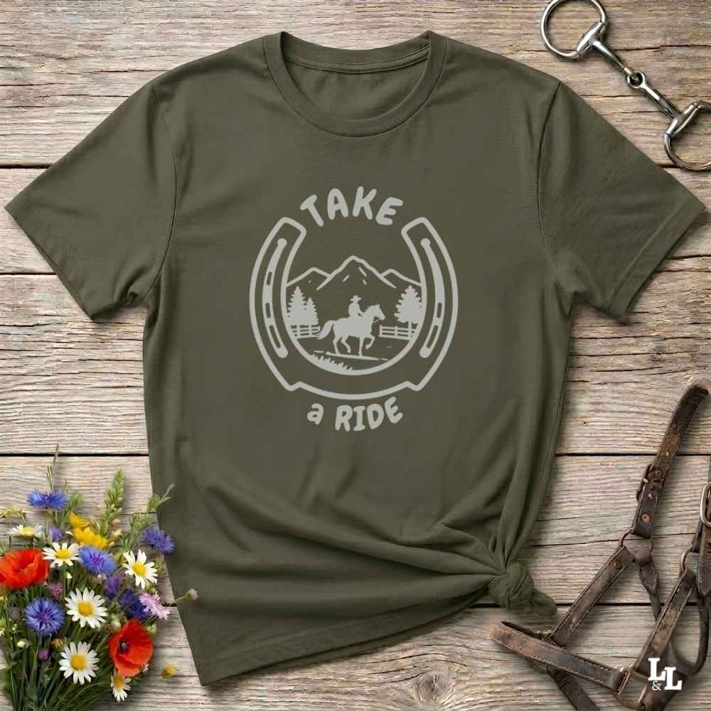 An olive green short-sleeve t-shirt with a white graphic print reading 'TAKE a RIDE' around a horse and mountain scene inside a horseshoe. The shirt is styled in a flat-lay on a rustic wooden surface with equestrian accessories and flowers.