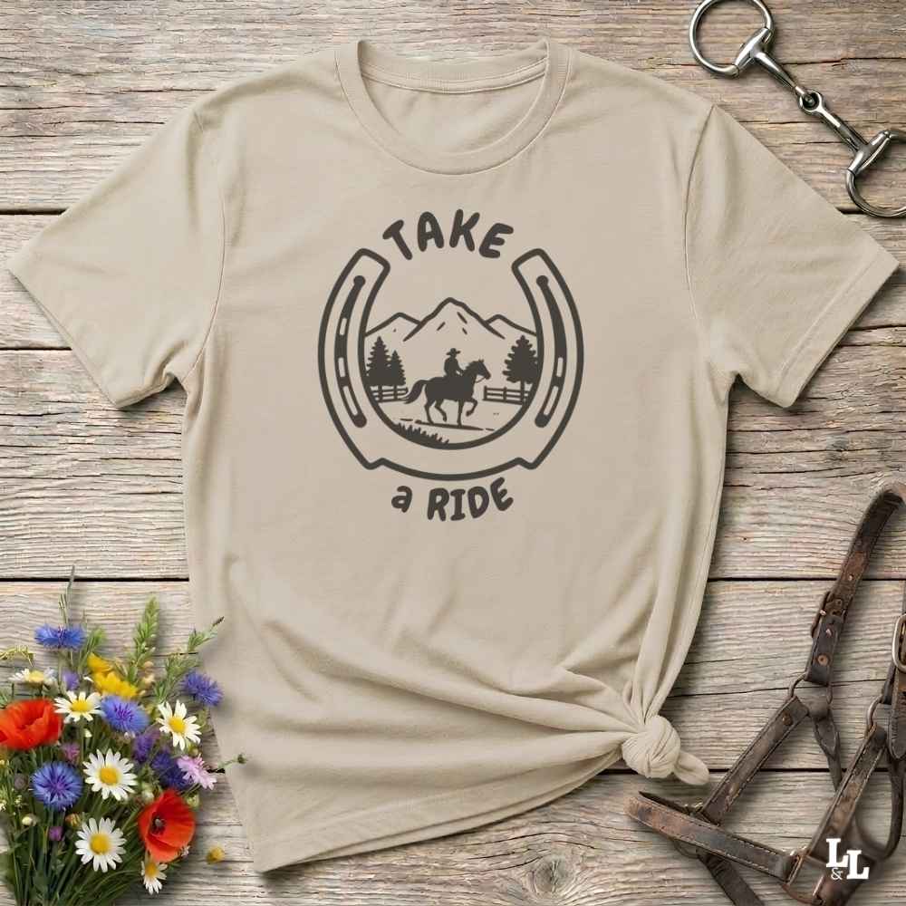 A beige graphic t-shirt with a black horseshoe and rider design reading 'TAKE a RIDE', styled flat on a rustic wood background with flowers and equestrian accessories.