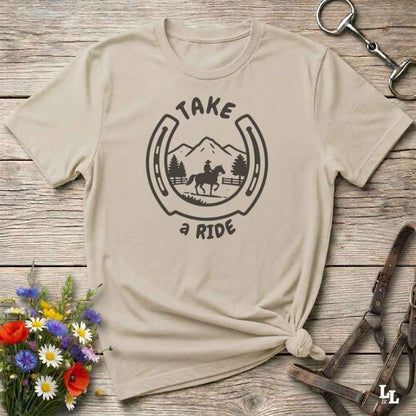 A beige graphic t-shirt with a black horseshoe and rider design reading 'TAKE a RIDE', styled flat on a rustic wood background with flowers and equestrian accessories.