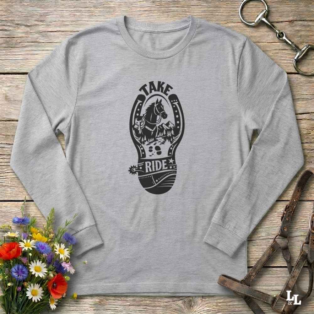 A light heather gray long-sleeve t-shirt with a black graphic print of a horse inside a horseshoe and the words 'TAKE RIDE'. The shirt is laid flat on a rustic wooden background with equestrian-themed props including a leather bridle, horse bit, and a bouquet of wildflowers.