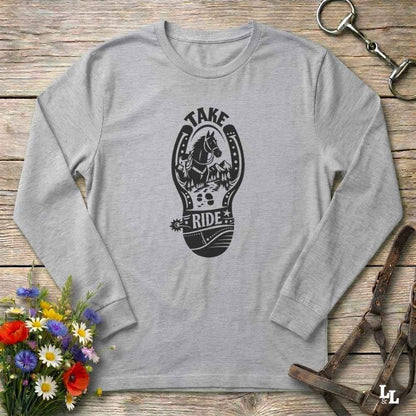 A light heather gray long-sleeve t-shirt with a black graphic print of a horse inside a horseshoe and the words 'TAKE RIDE'. The shirt is laid flat on a rustic wooden background with equestrian-themed props including a leather bridle, horse bit, and a bouquet of wildflowers.