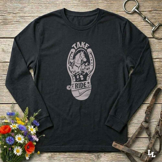 A black long-sleeve t-shirt with a gray graphic of a horse inside a horseshoe and the words 'TAKE RIDE', displayed flat on a rustic wooden background with equestrian and floral props.