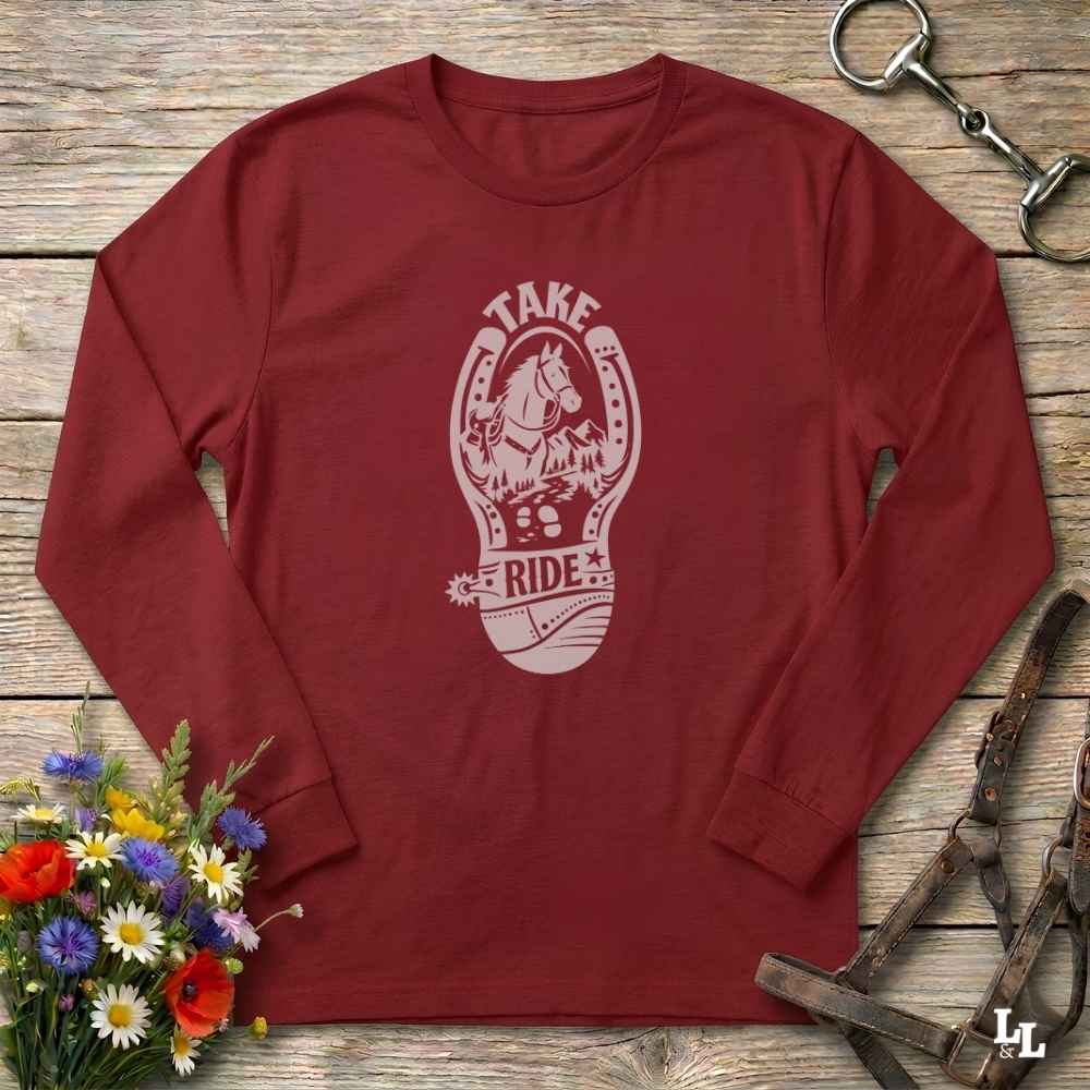 A long-sleeve crewneck t-shirt in maroon red, laid flat on a rustic wooden surface. The shirt features a light pink graphic in the center with a horse and rider inside a horseshoe and the words 'TAKE RIDE'.