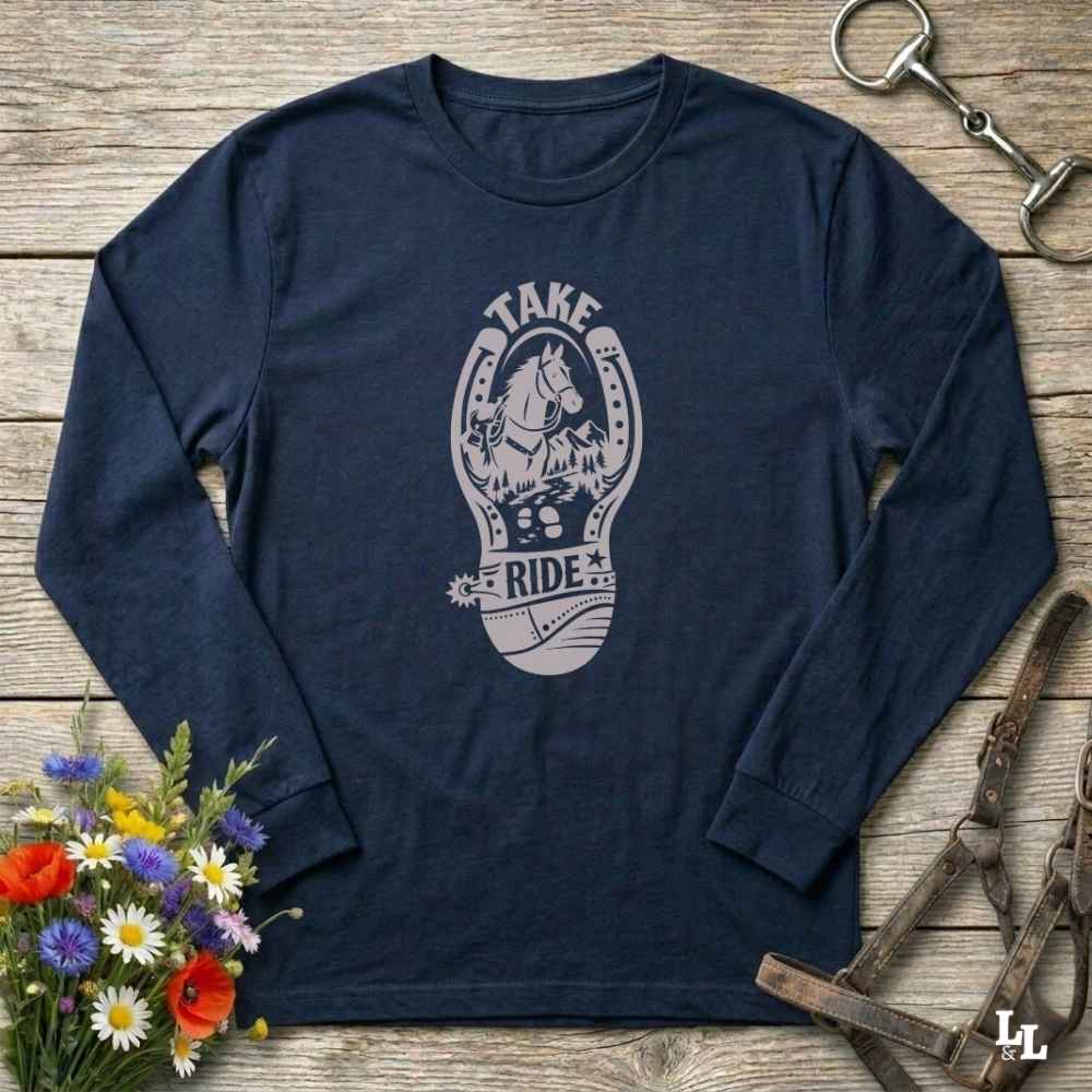 A navy blue long-sleeve t-shirt with a light gray graphic of a horse inside a horseshoe and the words 'TAKE RIDE', laid flat on a rustic wooden surface with equestrian accessories and flowers around it.