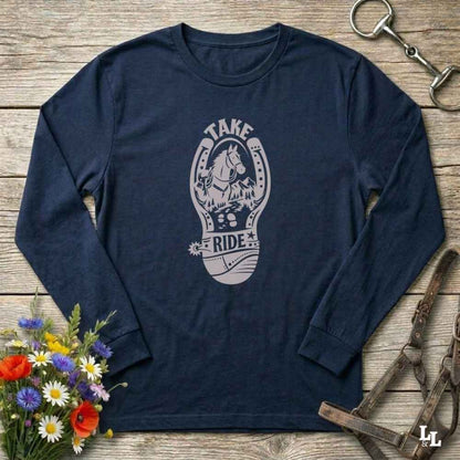 A navy blue long-sleeve t-shirt with a light gray graphic of a horse inside a horseshoe and the words 'TAKE RIDE', laid flat on a rustic wooden surface with equestrian accessories and flowers around it.