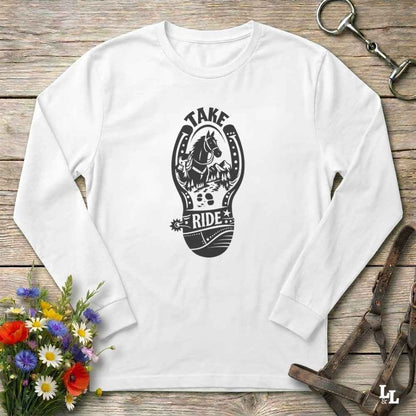 A white long-sleeve t-shirt with a black graphic print of a horse in a horseshoe and the words 'TAKE RIDE', displayed flat on a rustic wooden background with equestrian accessories and flowers.