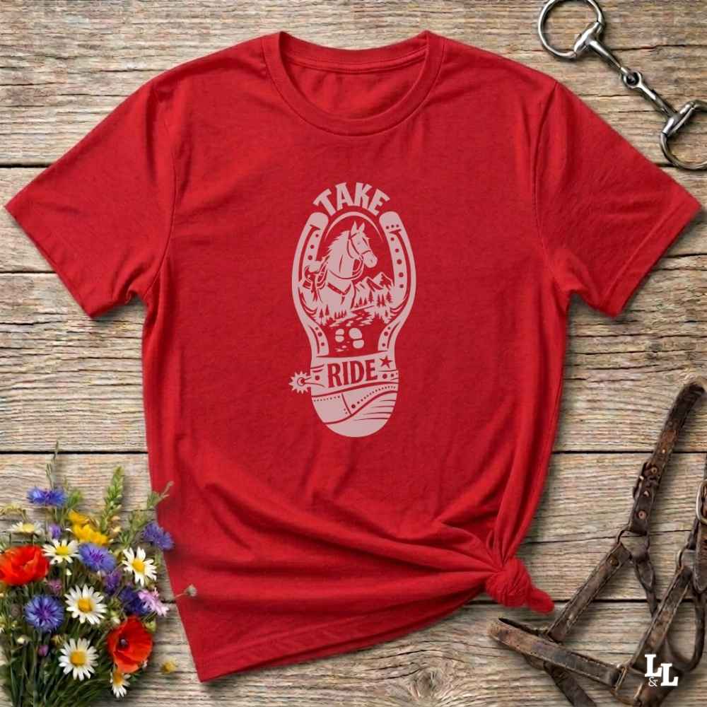A red short-sleeve graphic T-shirt laid flat on a rustic wooden surface, with a white horseshoe design featuring a horse and the words “TAKE RIDE.” Decorative flowers and equestrian tack are placed around the shirt.