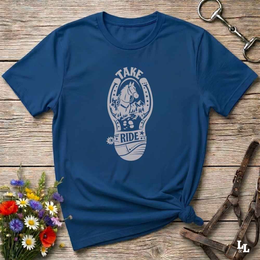 A navy blue short-sleeve t-shirt with a white graphic print of a horse inside a horseshoe and the words 'TAKE RIDE', styled on a rustic wooden surface with equestrian accessories and flowers.