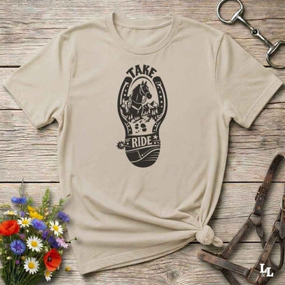 A beige short-sleeve t-shirt with a black graphic print of a horse and the words 'TAKE RIDE' inside a horseshoe design, styled on a rustic wooden background with equestrian accessories and flowers.