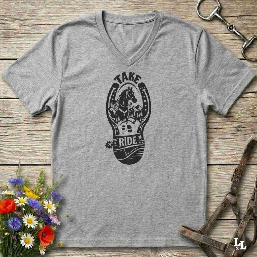 A gray V-neck t-shirt with a light gray graphic of a horse inside a horseshoe and the words 'TAKE RIDE', displayed on a rustic wooden background with equestrian accessories and flowers.
