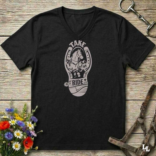A blackV-neck t-shirt with a light gray graphic of a horse inside a horseshoe and the words 'TAKE RIDE', displayed on a rustic wooden background with equestrian accessories and flowers.