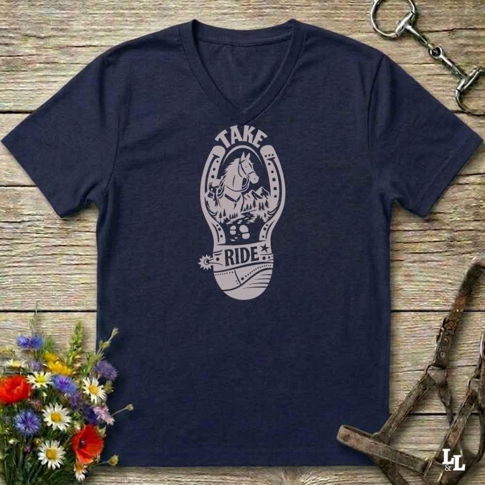 A navy blue V-neck t-shirt with a light gray graphic of a horse inside a horseshoe and the words 'TAKE RIDE', displayed on a rustic wooden background with equestrian accessories and flowers.