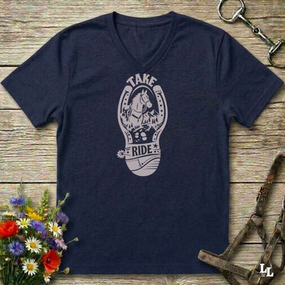 A navy blue V-neck t-shirt with a light gray graphic of a horse inside a horseshoe and the words 'TAKE RIDE', displayed on a rustic wooden background with equestrian accessories and flowers.