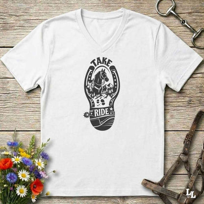 A white V-neck t-shirt with a light gray graphic of a horse inside a horseshoe and the words 'TAKE RIDE', displayed on a rustic wooden background with equestrian accessories and flowers.