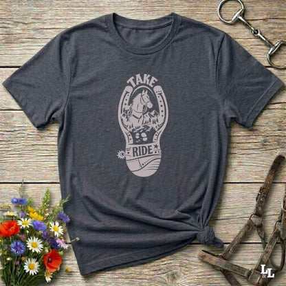 A dark heather gray t-shirt with a light gray graphic of a horse inside a horseshoe and the words 'TAKE RIDE', styled on a rustic wooden background with equestrian accessories and flowers.