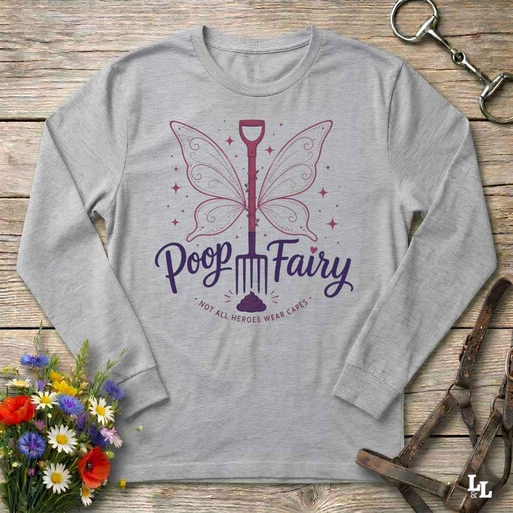 A light gray long-sleeve t-shirt with a 'Poop Fairy' graphic, laid flat on a rustic wooden background with decorative flowers and leather gear.