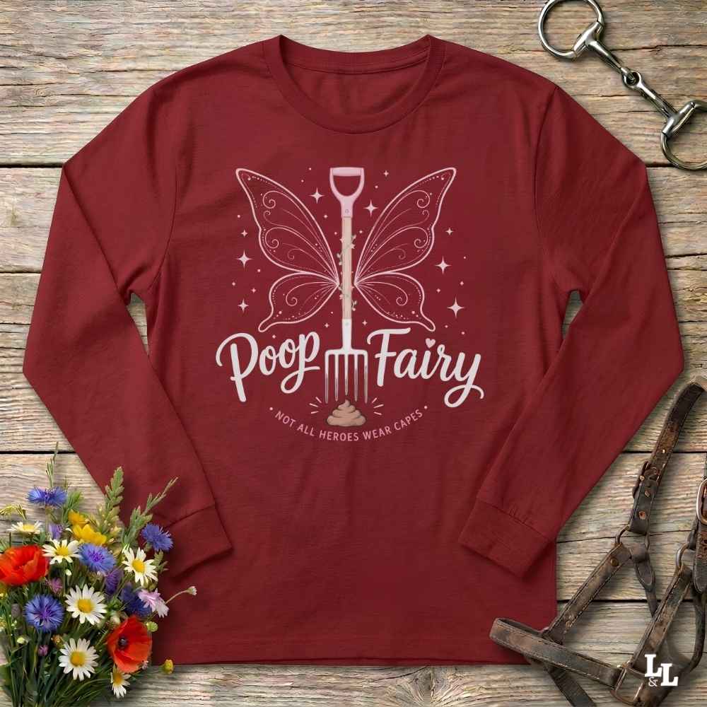 A long-sleeve crewneck t-shirt in maroon red, featuring a white graphic of a butterfly-shaped spade with the text 'Poop Fairy' and 'NOT ALL HEROES WEAR CAPES'. The shirt is displayed flat on a rustic wooden background with equestrian-themed props including a horse bit, leather bridle, and a small bouquet of wildflowers.