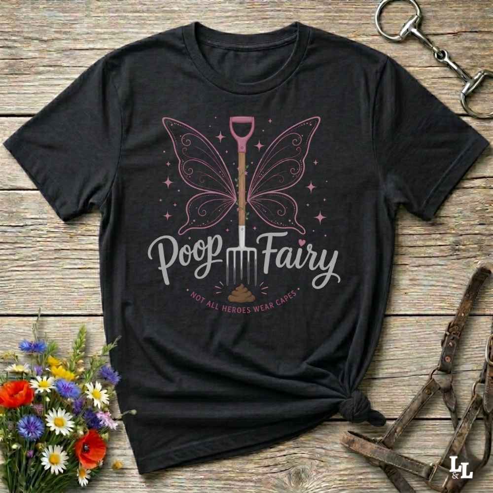 A black graphic t-shirt laid flat on a rustic wooden surface, styled with flowers, a horse bit, and a leather bridle. The shirt features a graphic of a butterfly-winged shovel with the text 'Poop Fairy' and 'NOT ALL HEROES WEAR CAPS'.