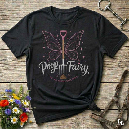 A black graphic t-shirt laid flat on a rustic wooden surface, styled with flowers, a horse bit, and a leather bridle. The shirt features a graphic of a butterfly-winged shovel with the text 'Poop Fairy' and 'NOT ALL HEROES WEAR CAPS'.