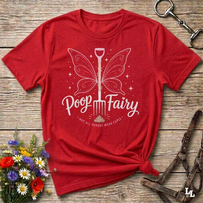 A red short-sleeve graphic T-shirt laid flat on a rustic wooden surface, with a white fairy-shovel graphic and the words 'Poop Fairy' and 'NOT ALL HEROES WERE CAVES.' The shirt is styled with flowers, a horse bit, and a leather bridle around it.