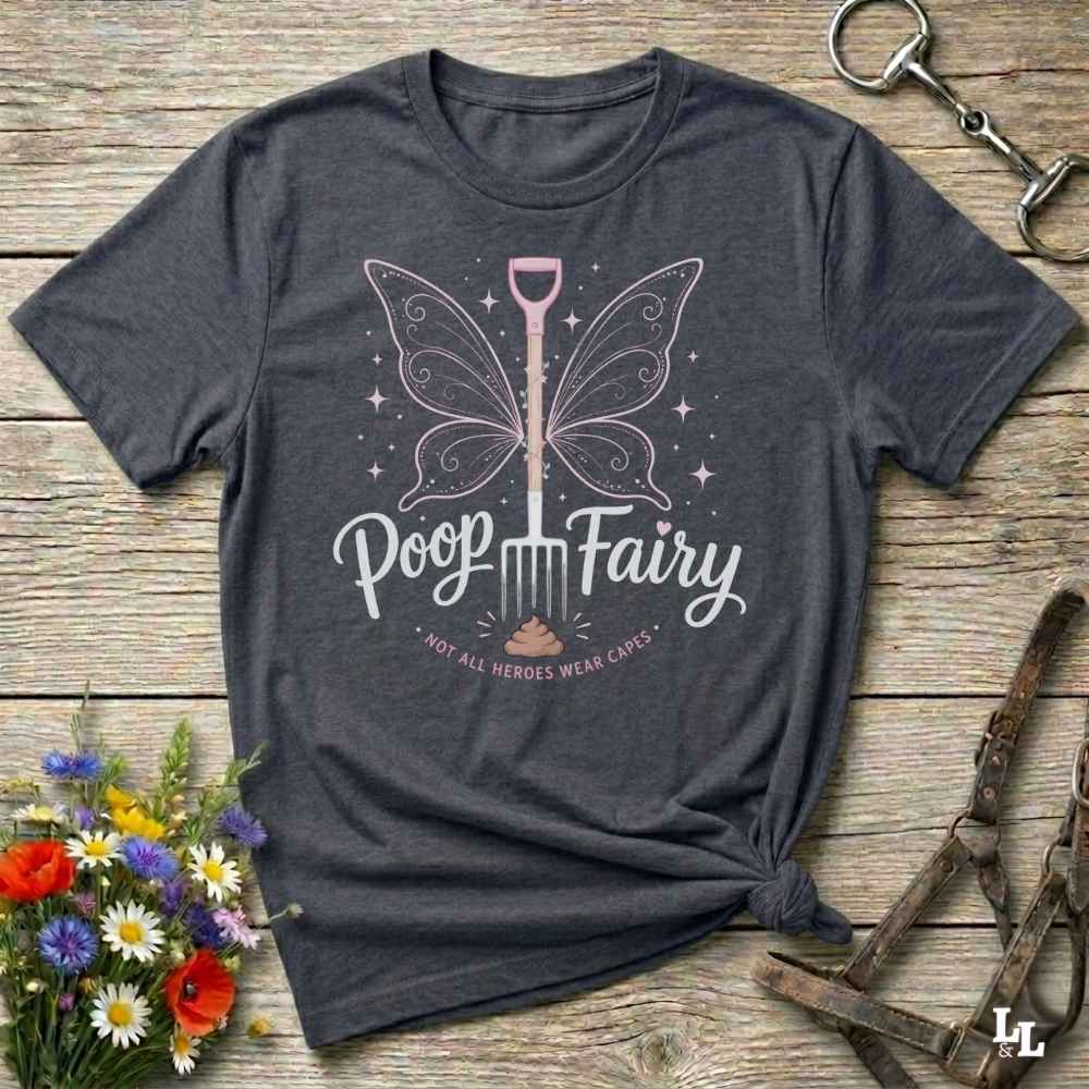 A dark charcoal gray graphic t-shirt laid flat on a rustic wooden surface, styled with a bouquet of flowers, a horse bit, and a leather bridle. The t-shirt features a graphic of a butterfly-winged shovel with the text 'Poop Fairy' and 'I NOT ALL HEROES WERE CARE'.