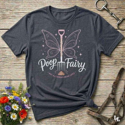 A dark charcoal gray graphic t-shirt laid flat on a rustic wooden surface, styled with a bouquet of flowers, a horse bit, and a leather bridle. The t-shirt features a graphic of a butterfly-winged shovel with the text 'Poop Fairy' and 'I NOT ALL HEROES WERE CARE'.