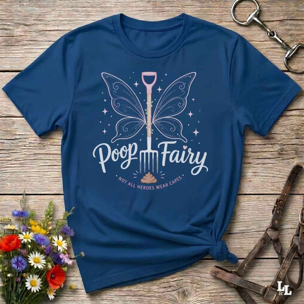 A navy blue graphic t-shirt laid flat on a rustic wooden surface, styled with a bouquet of flowers, a horse bit, and a leather bridle. The t-shirt features a graphic of a butterfly-winged shovel with the text 'Poop Fairy' and the subtitle '*NOT ALL HEROES WERE CAVES*'.