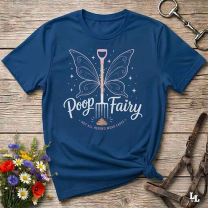 A navy blue graphic t-shirt laid flat on a rustic wooden surface, styled with a bouquet of flowers, a horse bit, and a leather bridle. The t-shirt features a graphic of a butterfly-winged shovel with the text 'Poop Fairy' and the subtitle '*NOT ALL HEROES WERE CAVES*'.