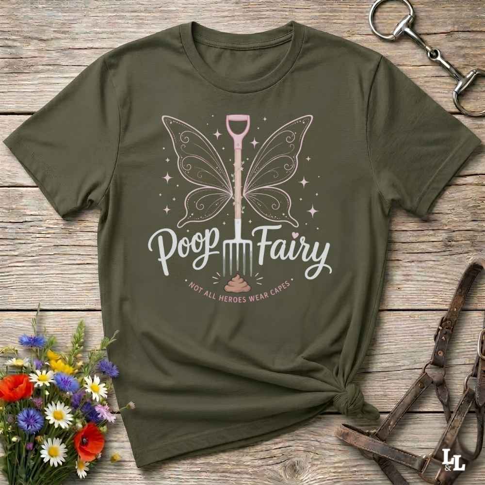 An olive green graphic t-shirt laid flat on a rustic wooden surface, styled with equestrian tack and a small bouquet of wildflowers. The shirt features a graphic of a butterfly-winged shovel with the text 'Poop Fairy' and the subtitle 'NOT ALL HEROES WEAR CAPES'.