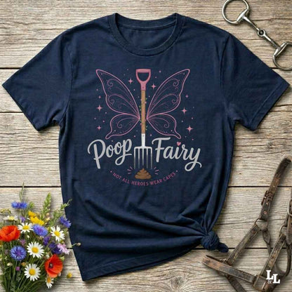 A navy blue graphic t-shirt laid flat on a rustic wooden surface, styled with flowers, horse tack, and a leather strap. The shirt features a graphic of a butterfly wings over a shovel with the text 'Poop Fairy' and 'NOT ALL HEROES WEAR CAPS'.