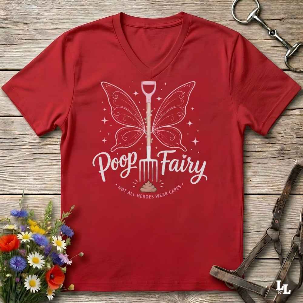 A red V-neck t-shirt with a white graphic of a butterfly-shaped shovel and the text 'Poop Fairy - NOT ALL HEROES WEAR CAES' laid flat on a rustic wooden background with horse tack and flowers as props.
