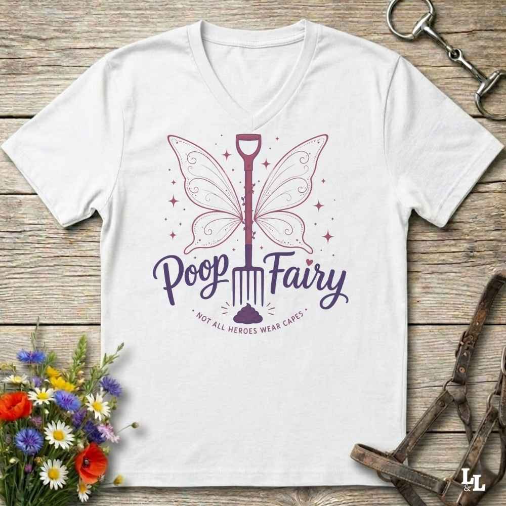 A white V-neck t-shirt with a purple graphic of a poop-shaped fairy and the text 'Poop Fairy - NOT ALL HEROES WEAR CAPES' laid flat on a rustic wooden background with equestrian accessories and flowers.