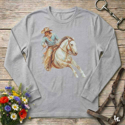 A light gray long-sleeve t-shirt with a vintage-style graphic of a cowboy riding a white horse, displayed on a rustic wooden background with decorative flowers and horse tack.