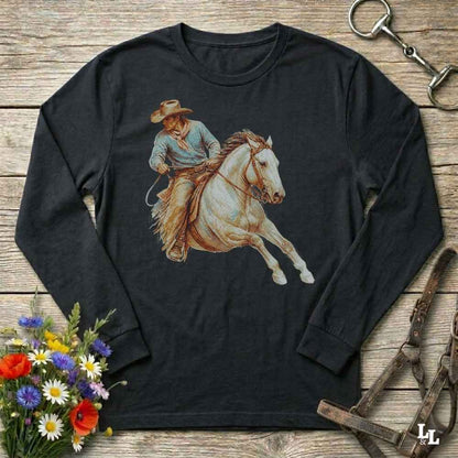 A black long-sleeve t-shirt with a western graphic of a cowboy riding a white horse, displayed on a rustic wooden background with decorative flowers and horse tack.