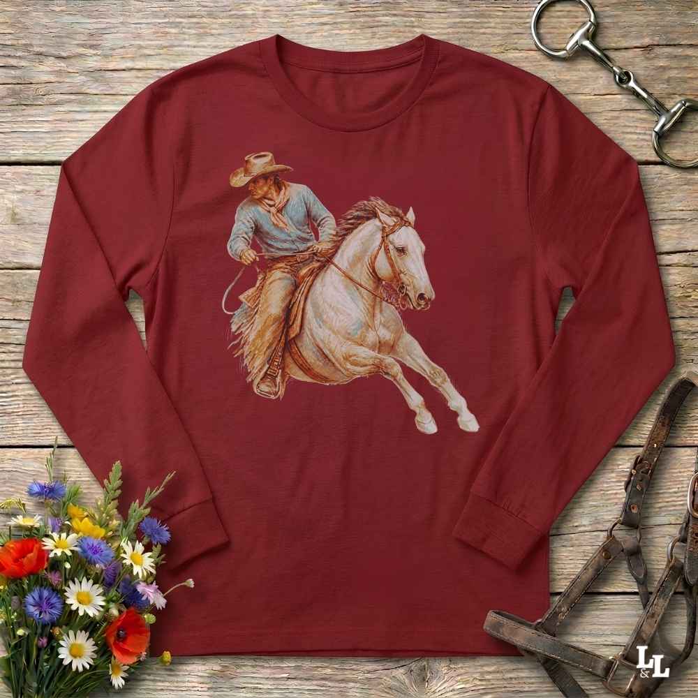 A long-sleeve crewneck t-shirt in maroon red, featuring a graphic print of a cowboy riding a white horse. The shirt is displayed in a flat-lay arrangement on a rustic wooden background, surrounded by equestrian tack, a small bouquet of wildflowers, and a silver horseshoe.