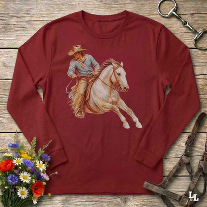 A long-sleeve crewneck t-shirt in maroon red, featuring a graphic print of a cowboy riding a white horse. The shirt is displayed in a flat-lay arrangement on a rustic wooden background, surrounded by equestrian tack, a small bouquet of wildflowers, and a silver horseshoe.