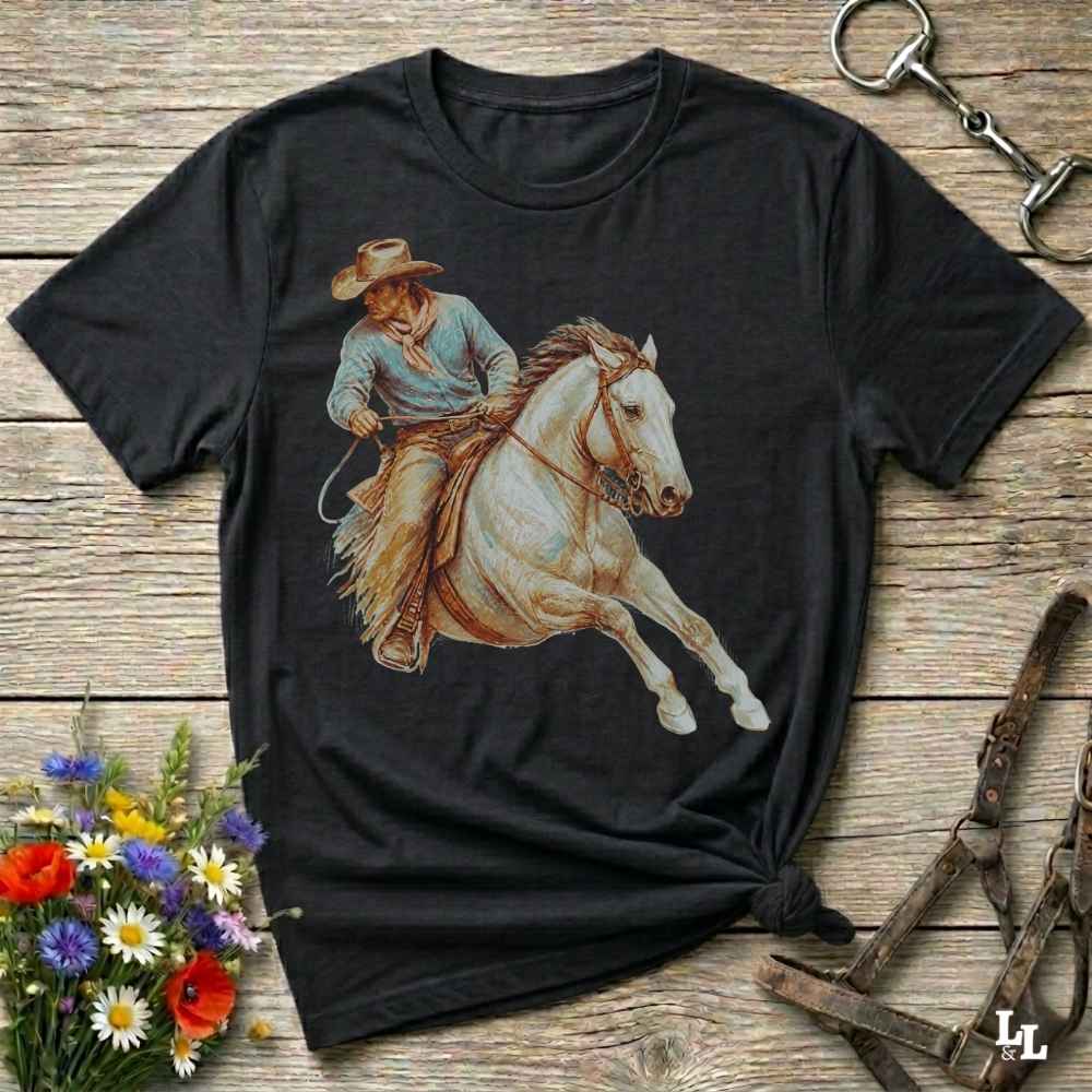 A black short-sleeve graphic t-shirt featuring a western cowboy riding a white horse, laid flat on a rustic wooden surface with floral and equestrian props.